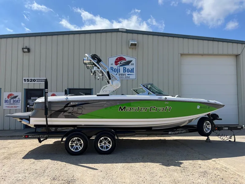 Slide: The Image of 2013 MasterCraft X-25 boat with green accents on a trailer, parked outside Boji Boat building. - 0