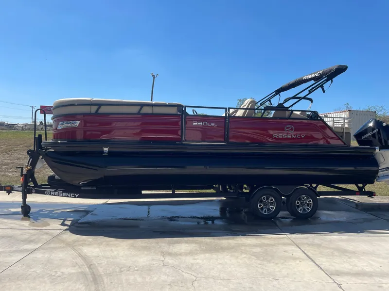 Slide: The Image of 2024 Regency 230 LE3 Sport pontoon boat on trailer, red and black design, parked outdoors. - 7