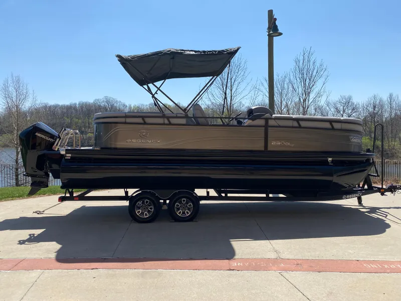 The Image of 2024 Regency 230 DL3 pontoon boat on trailer, parked outdoors under clear sky. - 0