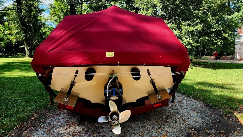 Slide: The Image of 1996 Crownline 266 BR boat with red cover, parked on gravel, surrounded by trees. - 13
