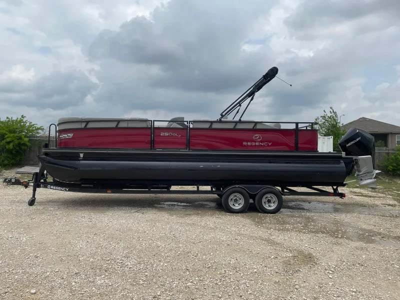 The Image of 2023 Regency 250DL3 pontoon boat on trailer, red and black design, parked outdoors. - 0