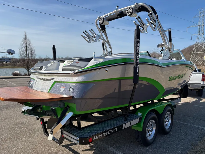 Slide: The Image of 2015 MasterCraft X23 boat on trailer, silver and green, parked near water. - 6