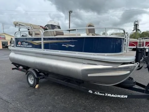 Slide: The Image of 2025 Sun Tracker Bass Buggy 16 XL Select pontoon boat on trailer, parked outdoors. - 2