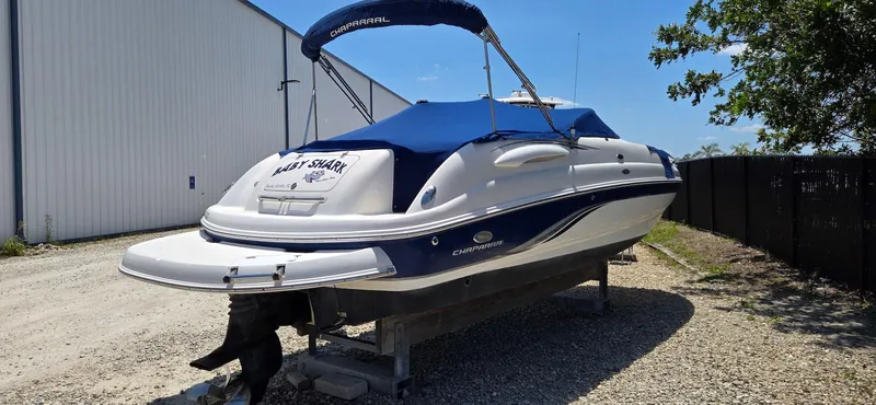 Slide: The Image of 2006 Chaparral Sunesta 254 boat with blue cover, parked on gravel near a building. - 2