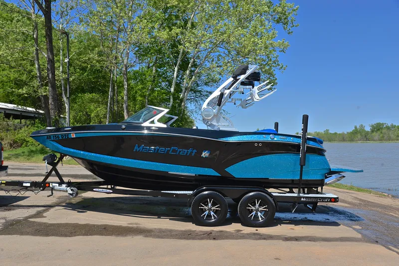 Slide: The Image of 2015 MasterCraft X20 boat on trailer by lakeside, featuring sleek black and blue design. - 11