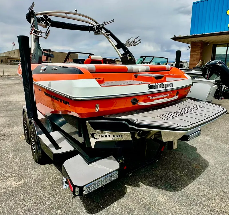 Slide: The Image of 2020 Malibu 21VLX boat in vibrant orange, parked outdoors with a wakeboard tower. - 2