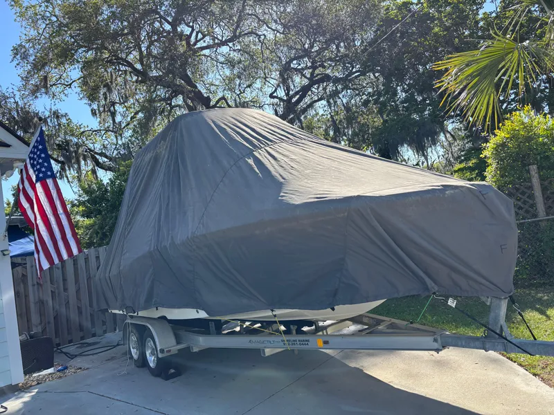 Slide: The Image of 2018 NauticStar 211 Hybrid boat covered on trailer, parked outdoors with American flag nearby. - 1