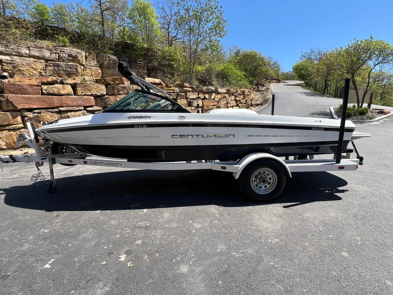 Slide: The Image of 2016 Centurion Carbon Pro boat on trailer, parked on a sunny road with stone wall backdrop. - 5