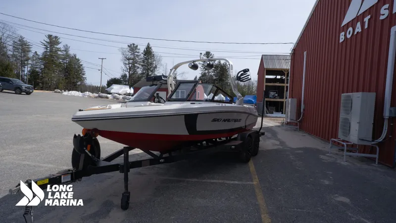Slide: The Image of 2025 Nautique 200 boat on trailer at Long Lake Marina, parked beside red building. - 4