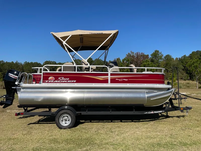 Slide: The Image of 2025 Sun Tracker Bass Buggy 18 DLX pontoon boat on trailer, parked on grass. - 2