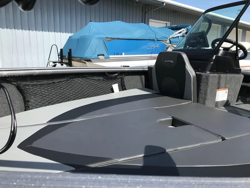 Slide: The Image of 2025 Lund 1875 Impact XS Sport boat interior with seating and storage, docked near blue-covered boat. - 13