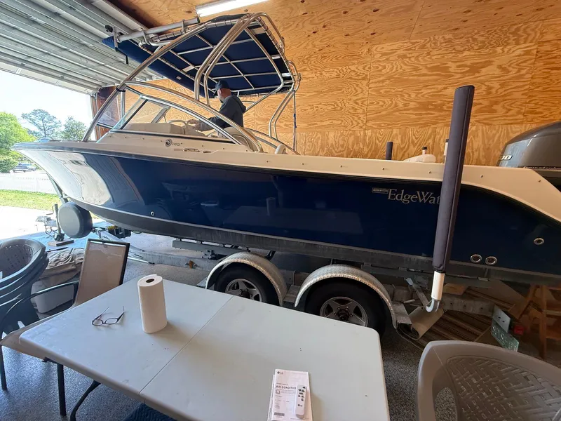 Slide: The Image of 2012 Edgewater 245 CX boat in garage, blue hull, on trailer, with table and chairs nearby. - 0