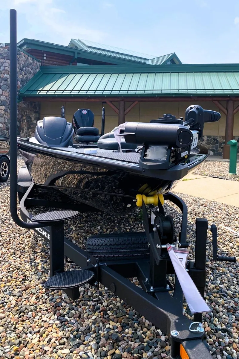 Slide: The Image of 2025 Tracker Savage 215 boat on trailer, parked on gravel near a building with green roof. - 2
