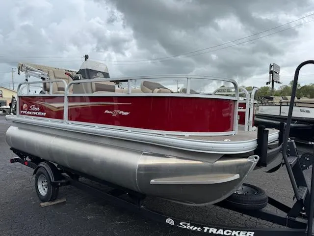 Slide: The Image of 2025 Sun Tracker Bass Buggy 16 XL Select pontoon boat on trailer, under cloudy sky. - 2