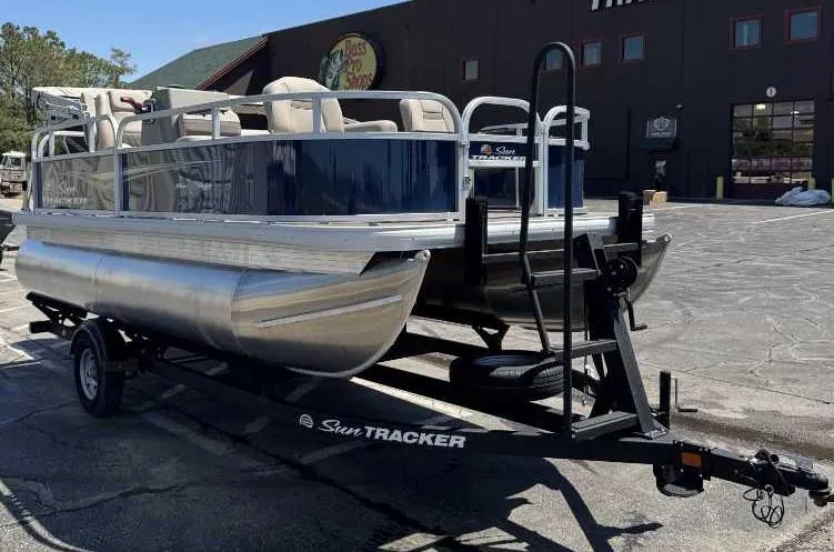 Slide: The Image of 2025 Sun Tracker Bass Buggy 16 XL Select pontoon boat on trailer, parked outdoors. - 2