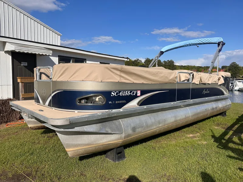 Slide: The Image of 2011 Avalon Windjammer 24' pontoon boat with cover, parked on grass near a building. - 1