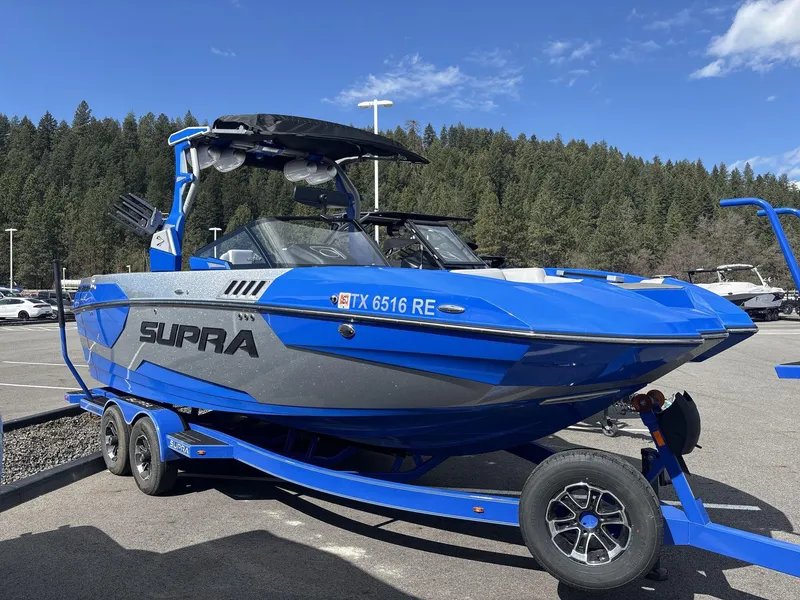 Slide: The Image of 2022 Supra SE 450 boat in blue on trailer, parked outdoors with forest background. - 1