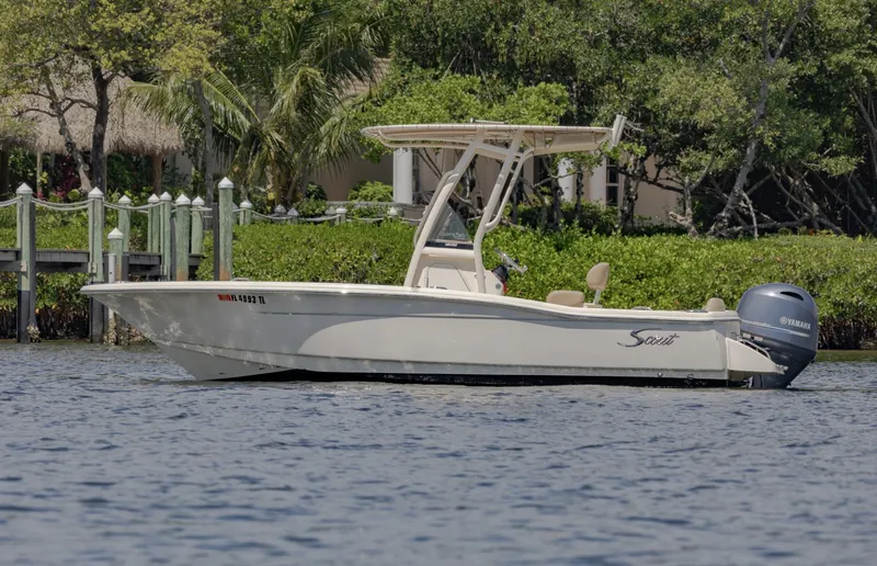 Slide: The Image of 2023 Scout 195 Sportfish boat on calm water near a lush, green shoreline. - 1
