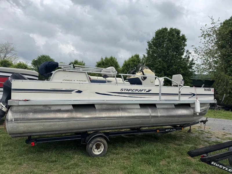 Slide: The Image of 2000 Starcraft 200 Classic pontoon boat on trailer, parked on grass under cloudy sky. - 13