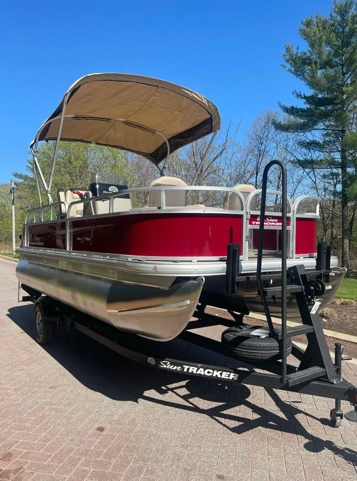 Slide: The Image of 2025 Sun Tracker Bass Buggy 18 DLX pontoon boat on trailer, red exterior, parked outdoors. - 2