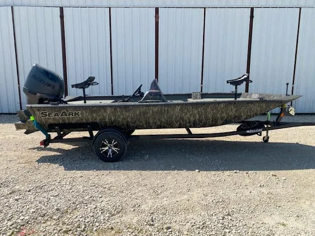 Slide: The Image of 2025 SeaArk Shadow 18 boat with Suzuki motor, parked on gravel near a white building. - 0