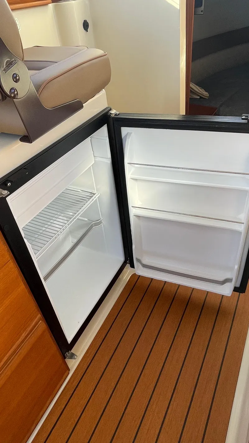 Slide: The Image of 2023 Ranger Tugs R-27 boat interior with open refrigerator on wooden flooring. - 27