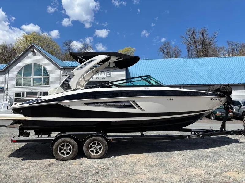 Slide: The Image of 2019 Regal 25 RX Surf boat on trailer, parked near a marina with blue roof. - 16