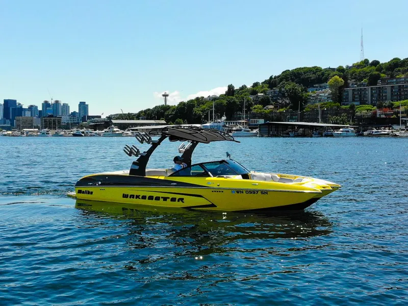 The Image of 2016 Malibu 24.5 MKZ boat on water, city skyline and greenery in background. - 1