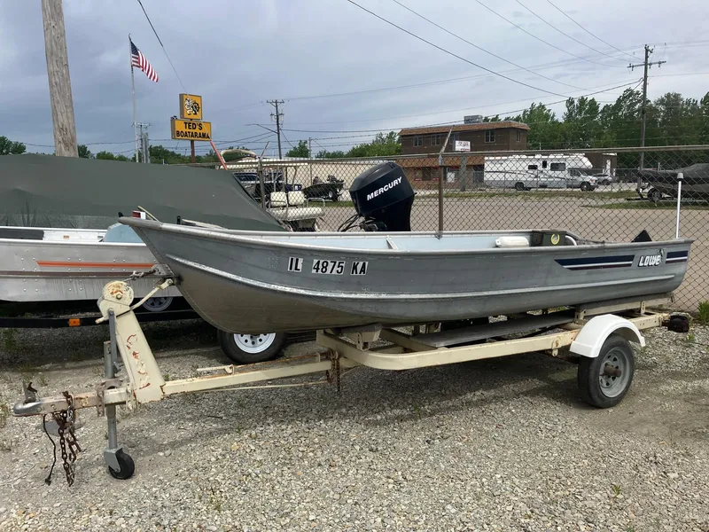 The Image of 1988 Lowe 14' V boat on trailer, parked outdoors near a fence and other vehicles. - 0