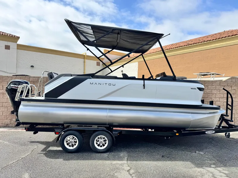 The Image of 2025 Manitou Cruise 20 Bench Twin Tube pontoon boat on a trailer, parked outdoors. - 0