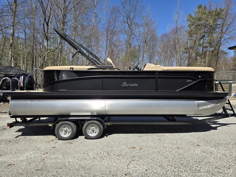 Slide: The Image of 2025 Barletta C22QC pontoon boat on trailer, parked outdoors, surrounded by trees. - 1
