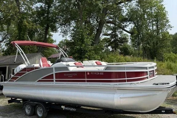 Slide: The Image of 2021 Bennington 24 RTSB pontoon boat with red accents on a trailer, surrounded by trees. - 0