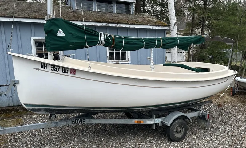 Slide: The Image of 2001 Com-Pac Picnic Cat sailboat on trailer, green sail cover, parked by blue building. - 0