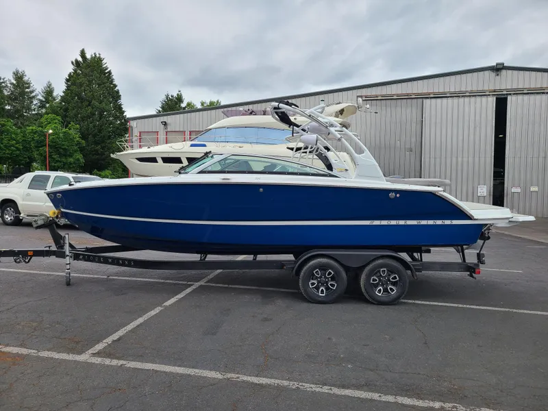 Slide: The Image of 2023 Four Winns H4 boat on trailer, blue hull, parked outdoors near warehouse. - 1