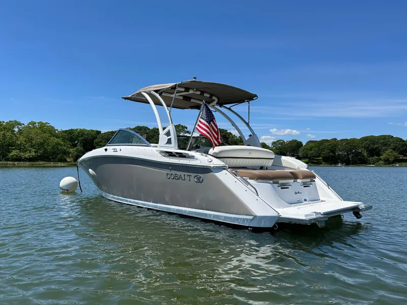 Slide: The Image of 2015 Cobalt 24SD boat on calm water with American flag, clear sky, and lush shoreline. - 0