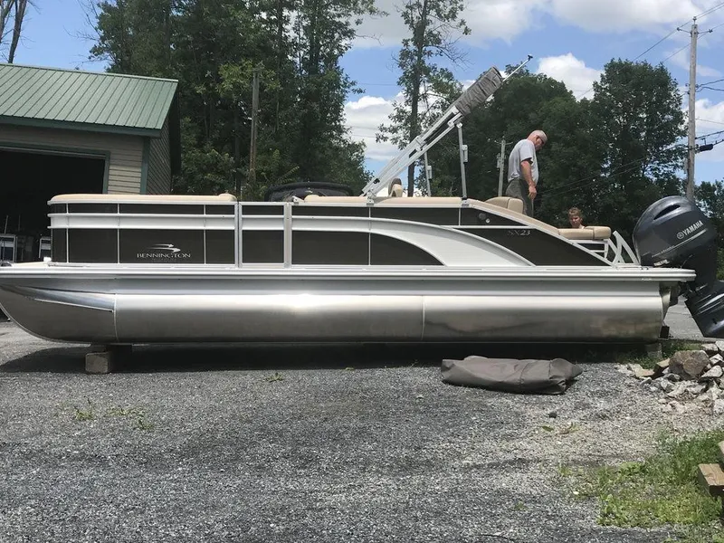 Slide: The Image of 2019 Bennington 23 SSBX pontoon boat on gravel, surrounded by trees and a building. - 1
