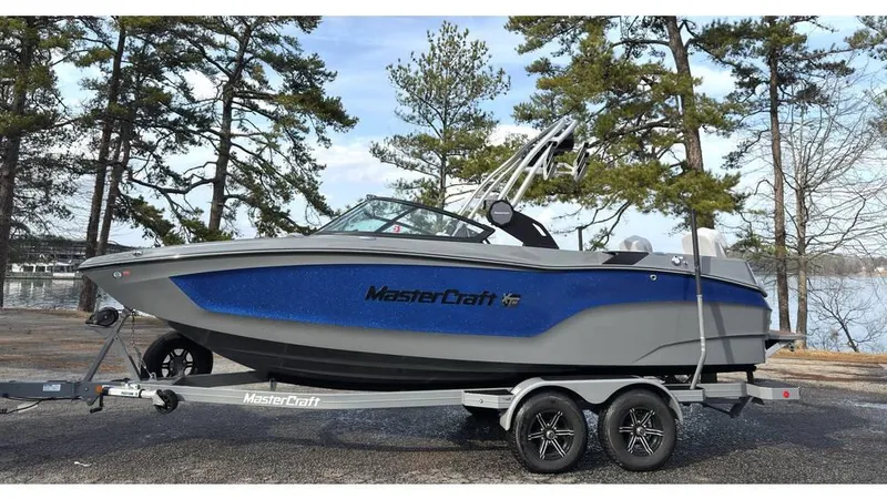 The Image of 2023 MasterCraft XT22 boat on trailer, parked near lake with trees in background. - 0