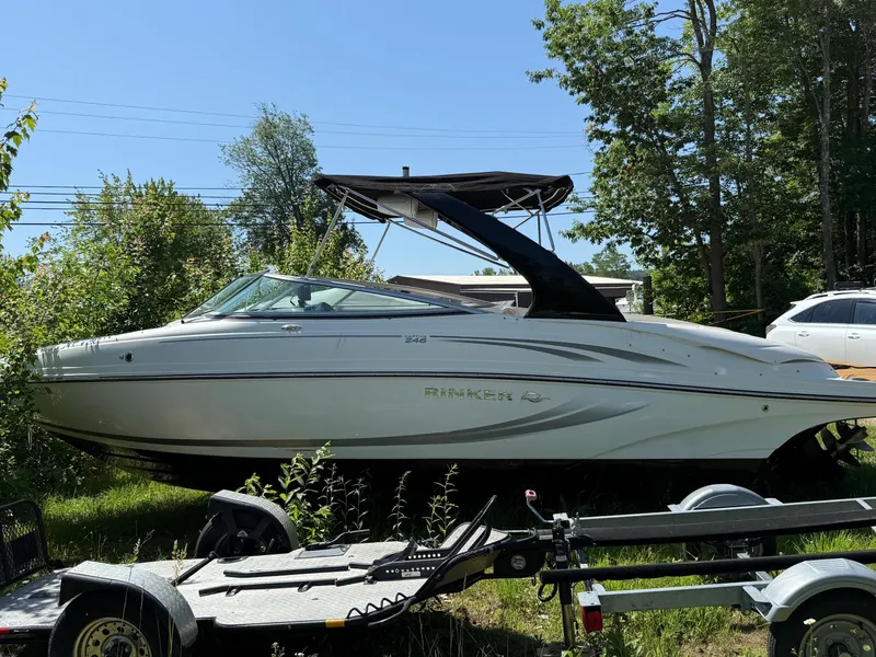 Slide: The Image of 2015 Rinker 246 Bowrider boat with canopy, parked on a trailer, surrounded by greenery. - 2