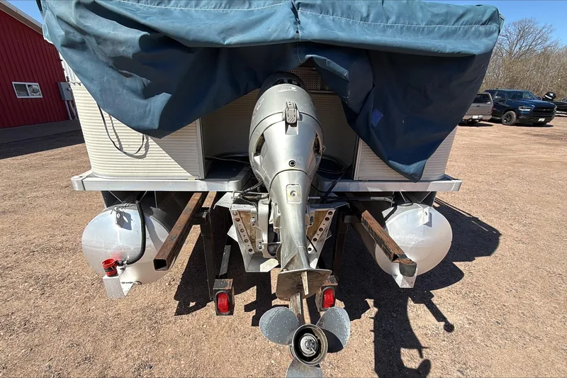 Slide: The Image of 1999 Smoker Craft Party Pontoon with outboard motor, covered and parked on a dirt surface. - 2