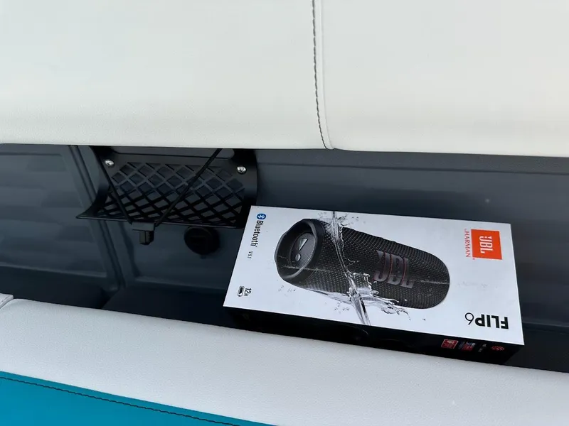 Slide: The Image of Boxed JBL Flip 6 speaker on Aura Quad Lounge 2025 seat with storage net. - 22
