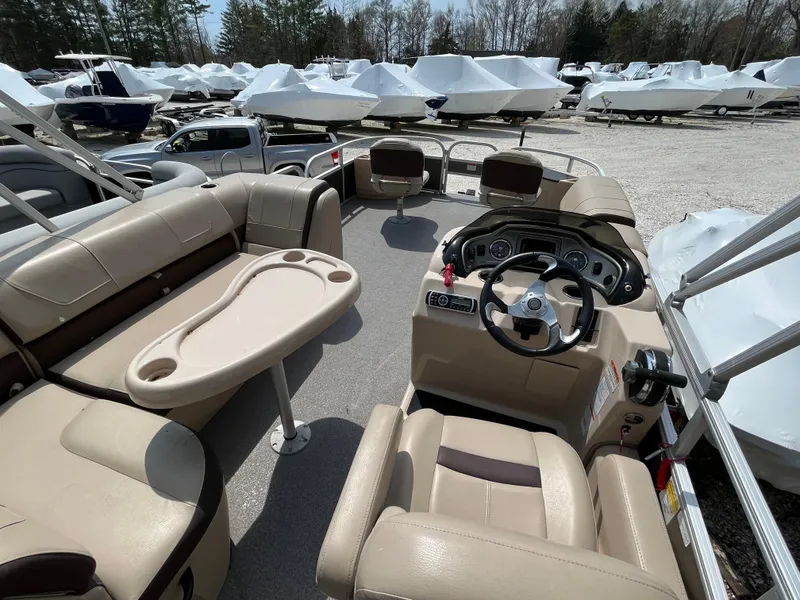 Slide: The Image of 2018 Sun Tracker Fishing Barge 22 interior with beige seating and control panel, parked among covered boats. - 19