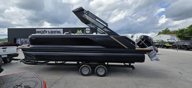 Slide: The Image of 2025 Manitou Explore pontoon boat on trailer, parked outdoors under cloudy sky. - 2