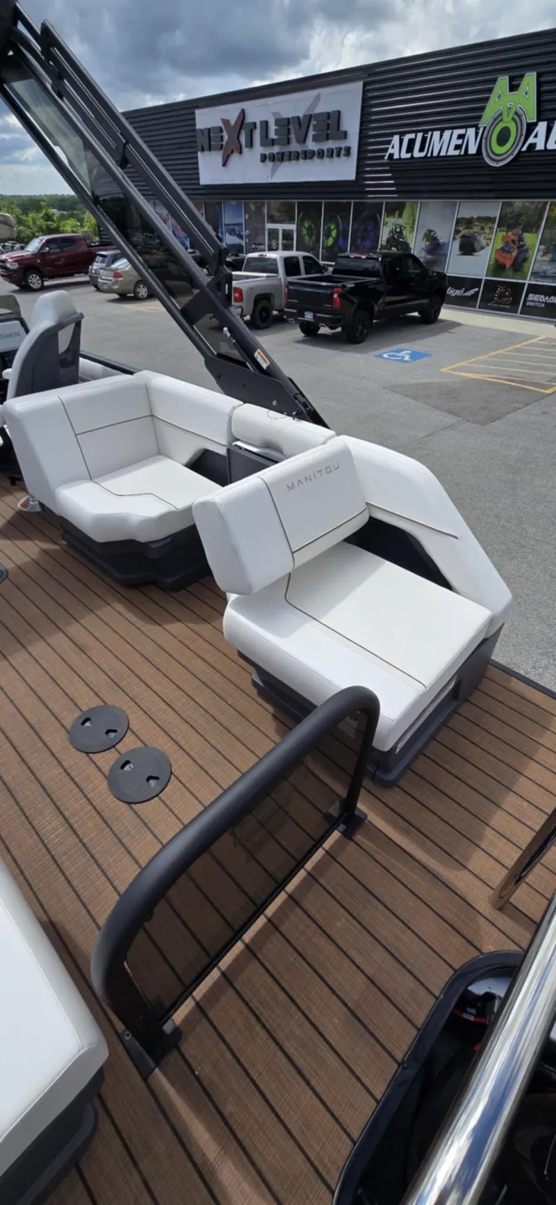 Slide: The Image of 2025 Manitou Explore boat with white seating and wooden deck, parked outside a dealership. - 23