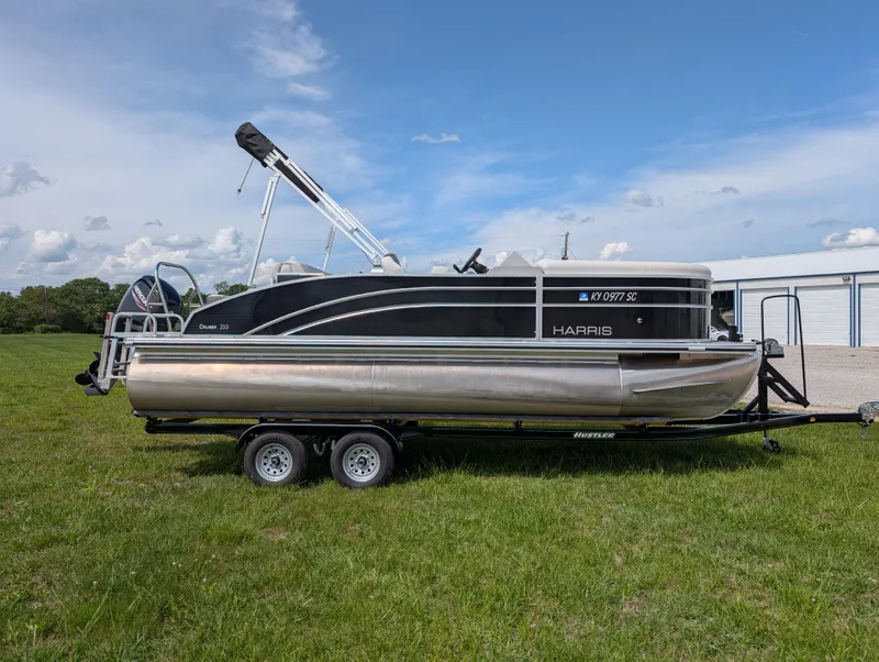 Slide: The Image of 2022 Harris Cruiser 210 pontoon boat on trailer, parked on grassy field under blue sky. - 1