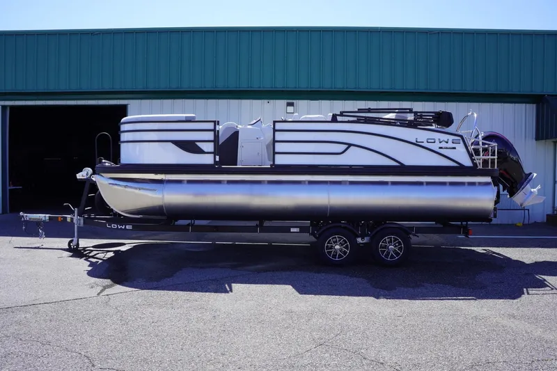 Slide: The Image of 2025 Lowe SS 210 DL pontoon boat on trailer, parked outside a warehouse. - 1