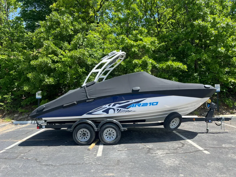 Slide: The Image of 2022 Yamaha AR210 boat on trailer, covered, parked in a lot with trees in the background. - 31