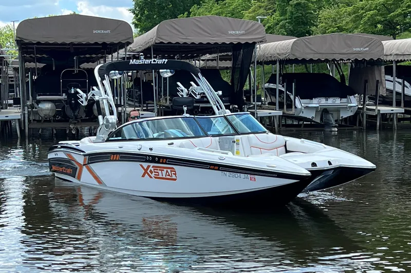 Slide: The Image of 2013 MasterCraft XStar boat docked on a calm lake, surrounded by covered slips. - 2