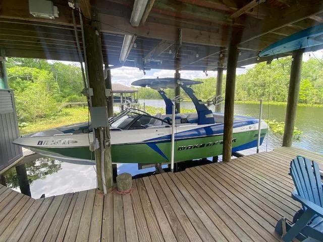 Slide: The Image of 2016 Malibu Wakesetter 25 LSV boat docked under a wooden shelter by a scenic river. - 1