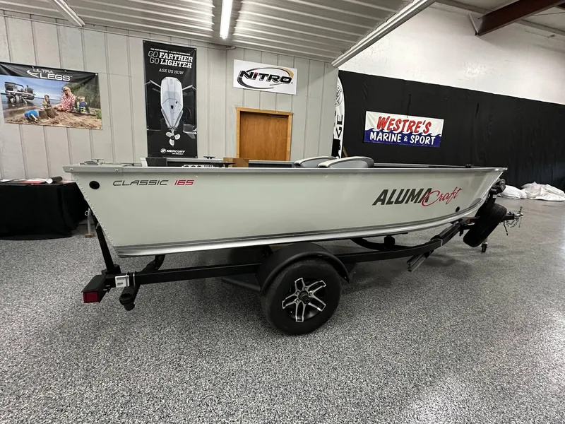 The Image of 2024 Alumacraft Classic 165 Tiller boat on trailer in showroom. - 0