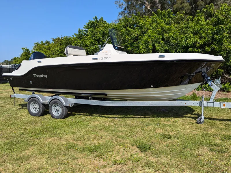 The Image of 2025 Bayliner Trophy T22CC boat on trailer, black hull, bimini top, parked on grass. - 0